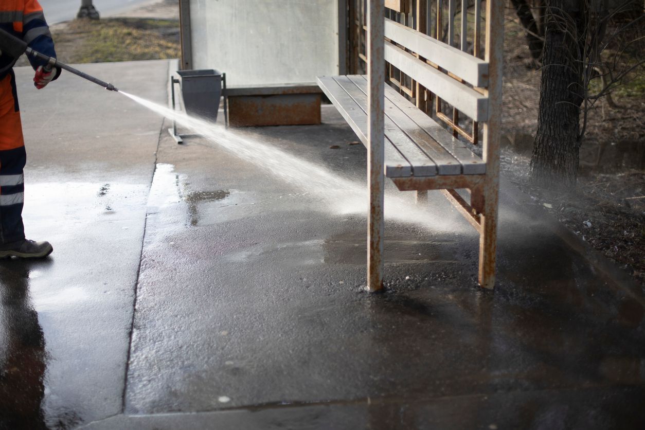 A worker in orange overalls uses a pressure washer to clean a sidewalk near a bus stop bench.