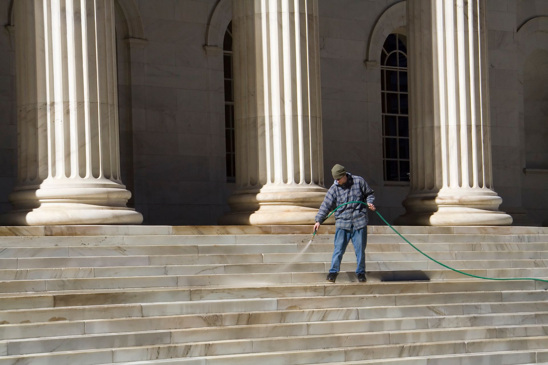Person washing steps of a building with large columns. Blue jeans, green hose, sunny day.