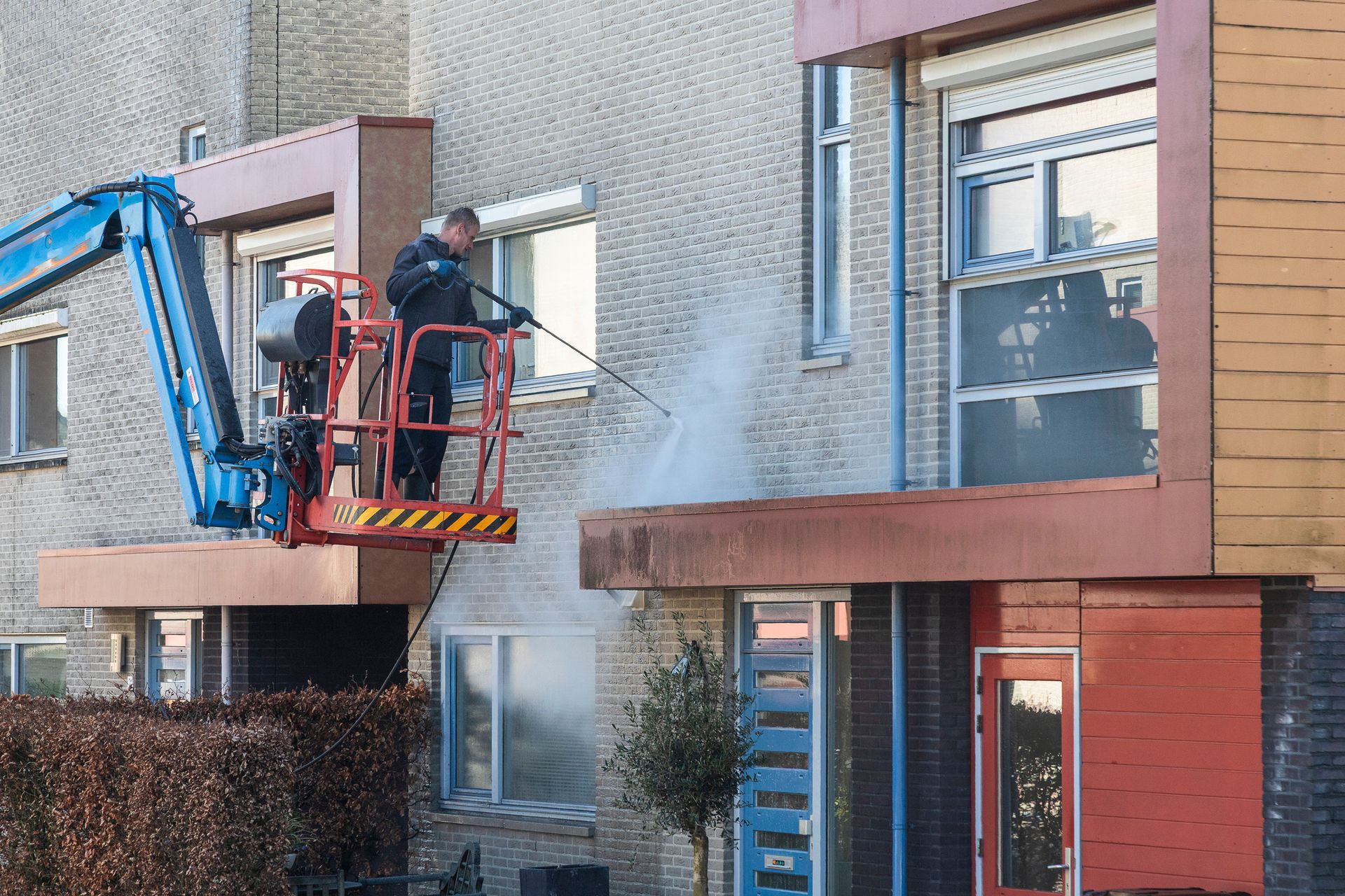 Man power washing a building's exterior from a blue lift; brick building with red accents.