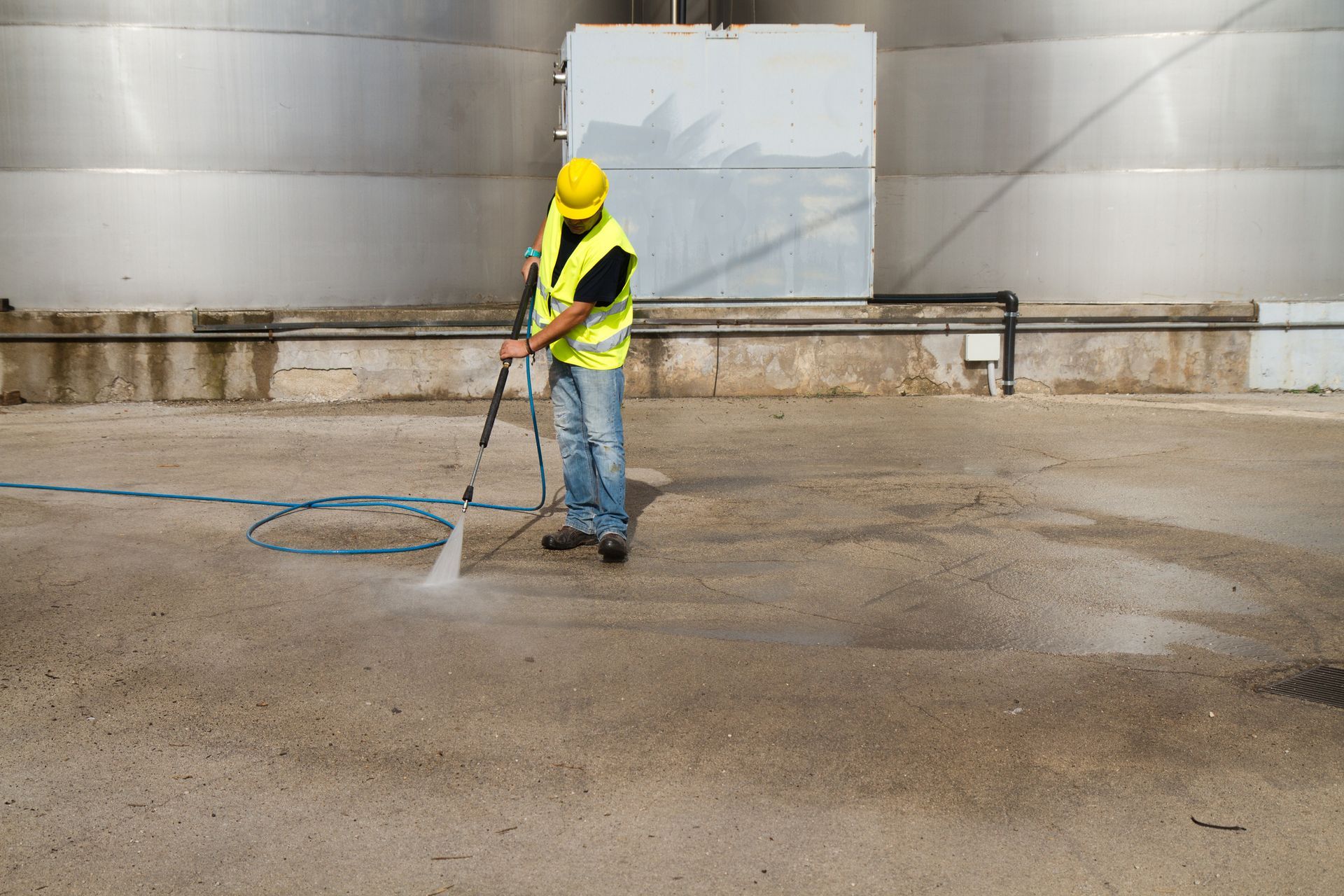 Worker in safety vest power washes a concrete industrial surface near large tanks.