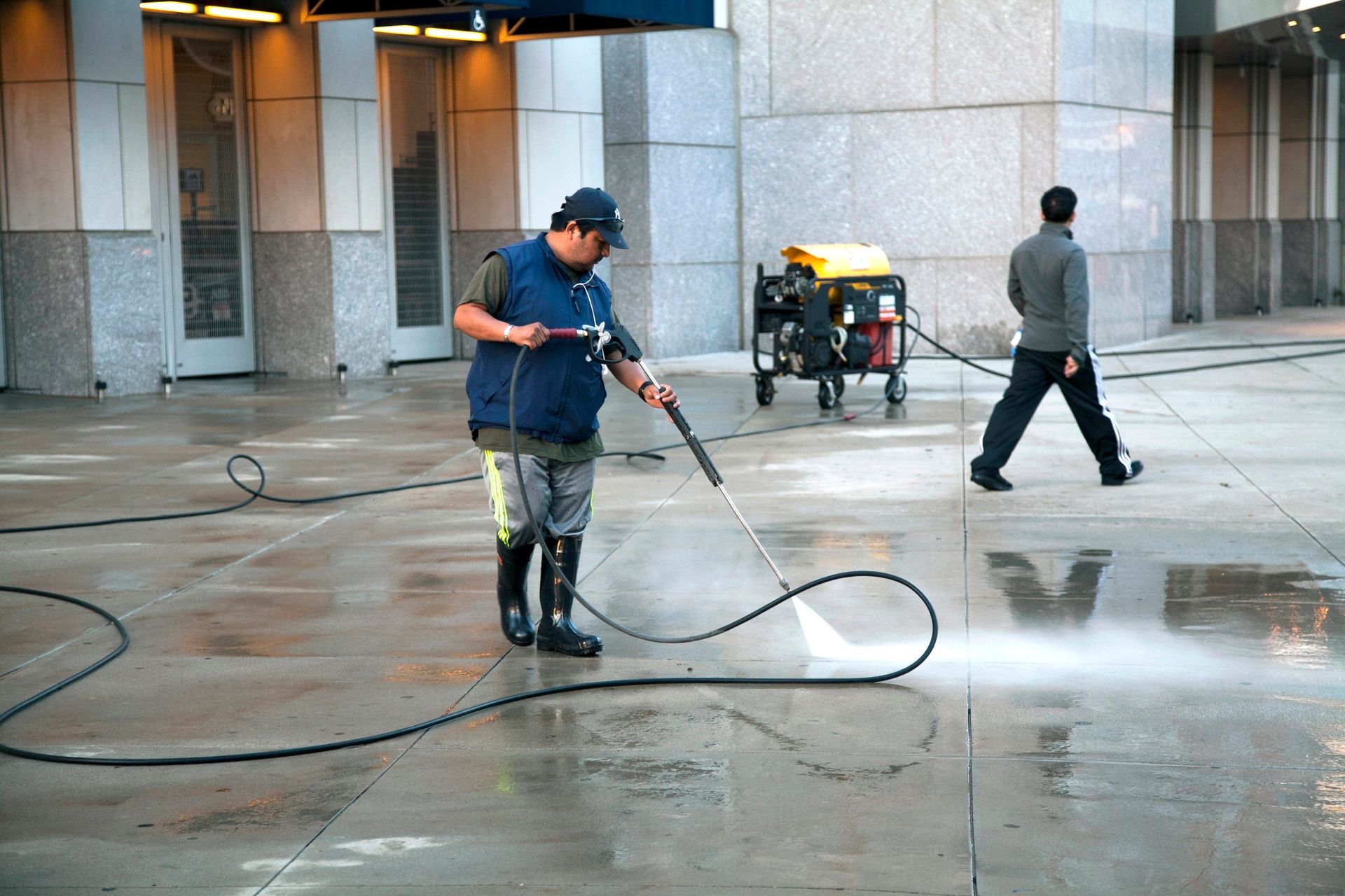 Man pressure washing a concrete sidewalk, equipment in the background, another person walks past.