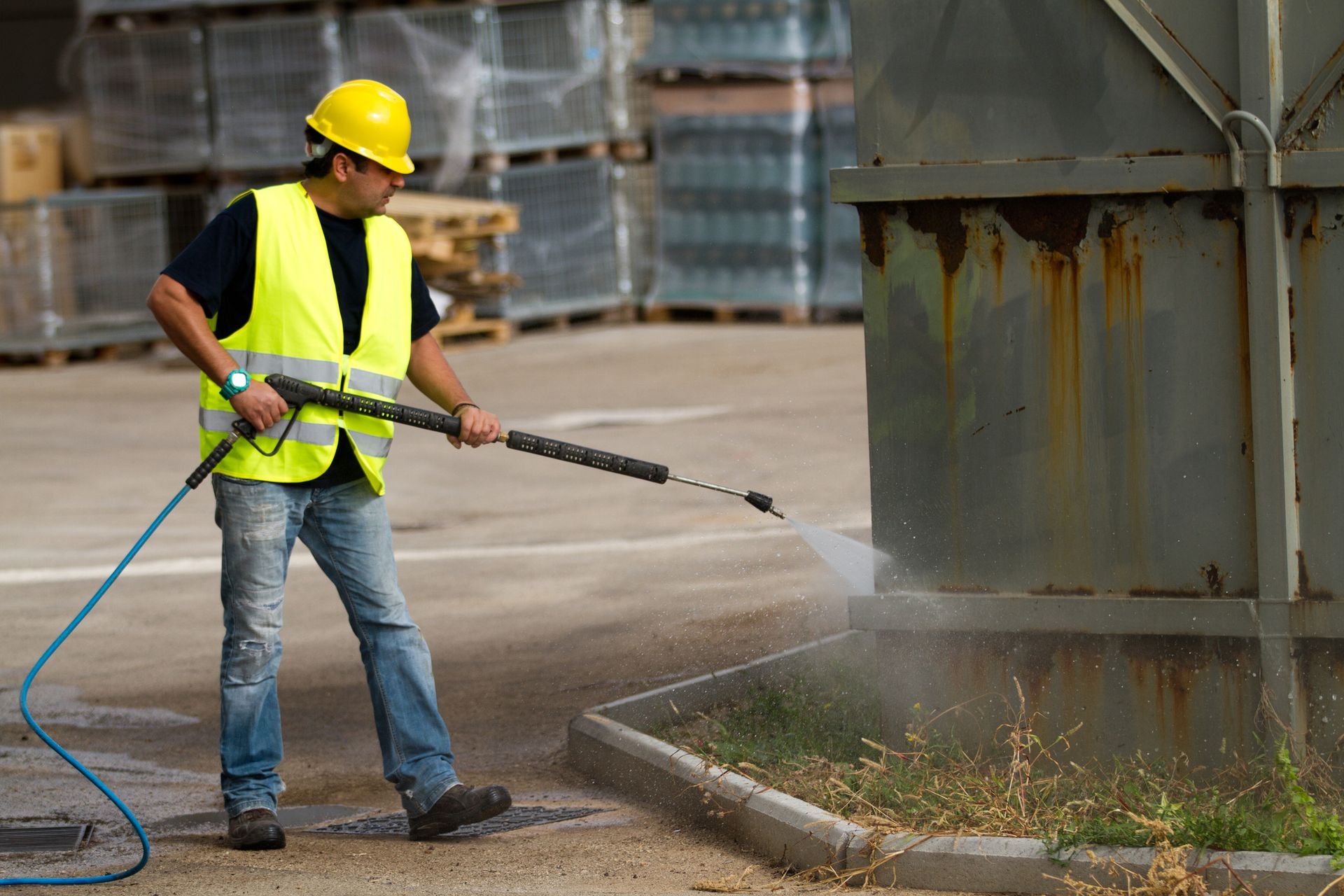 Man in safety gear pressure washing a concrete curb near a metal container.