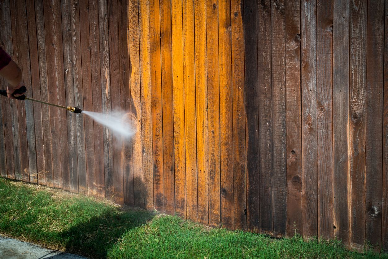 Pressure washer spraying water onto a weathered wooden fence, leaving a bright clean streak
