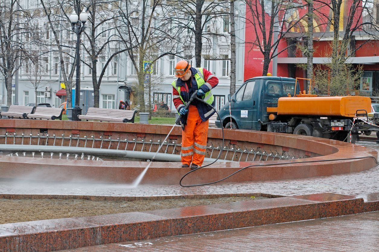 Worker in orange safety gear pressure-washes a wet stone fountain in a snowy city square.