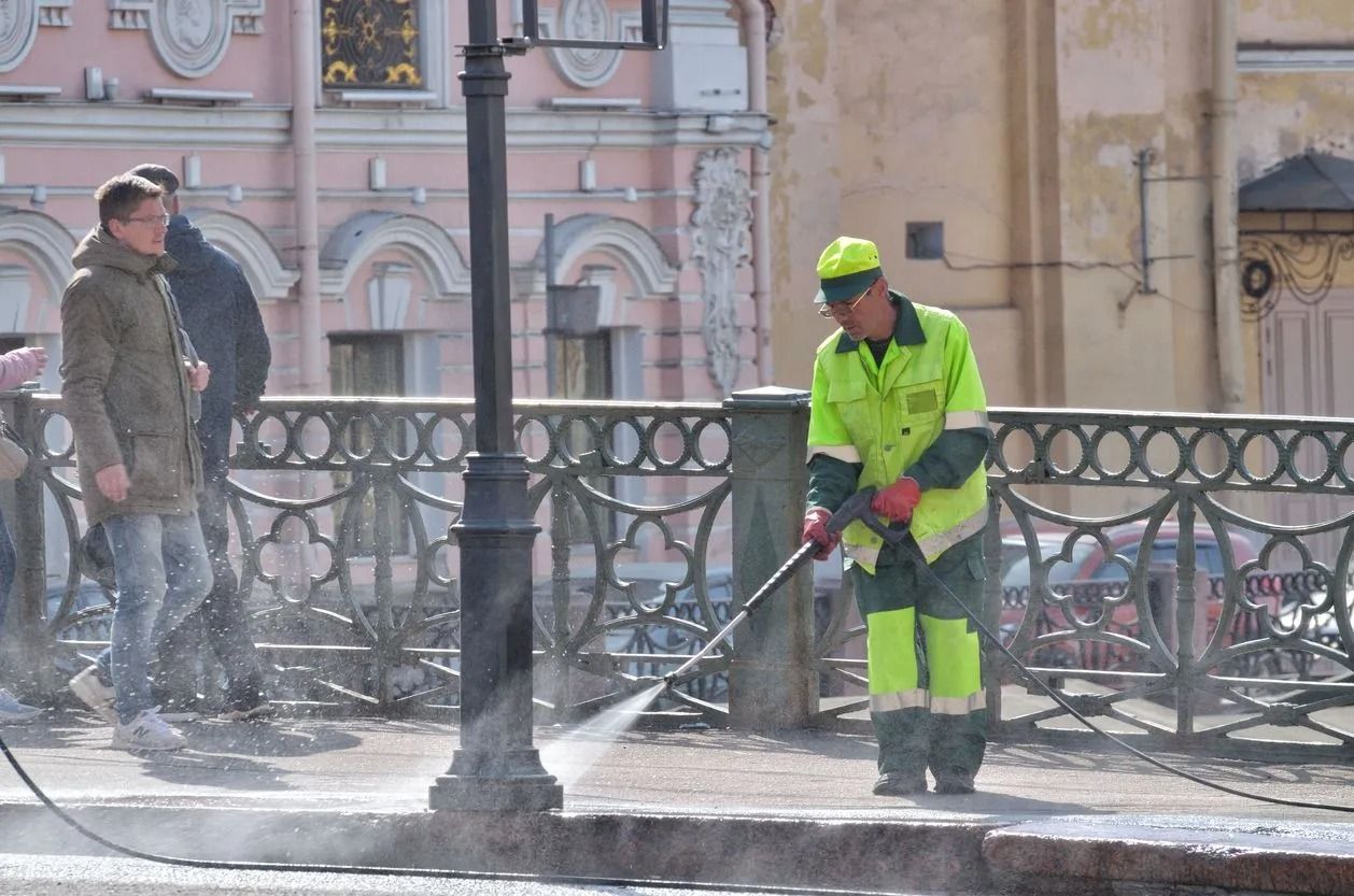 Worker in neon safety gear uses a hose to clear snow from a city sidewalk as pedestrians pass nearby