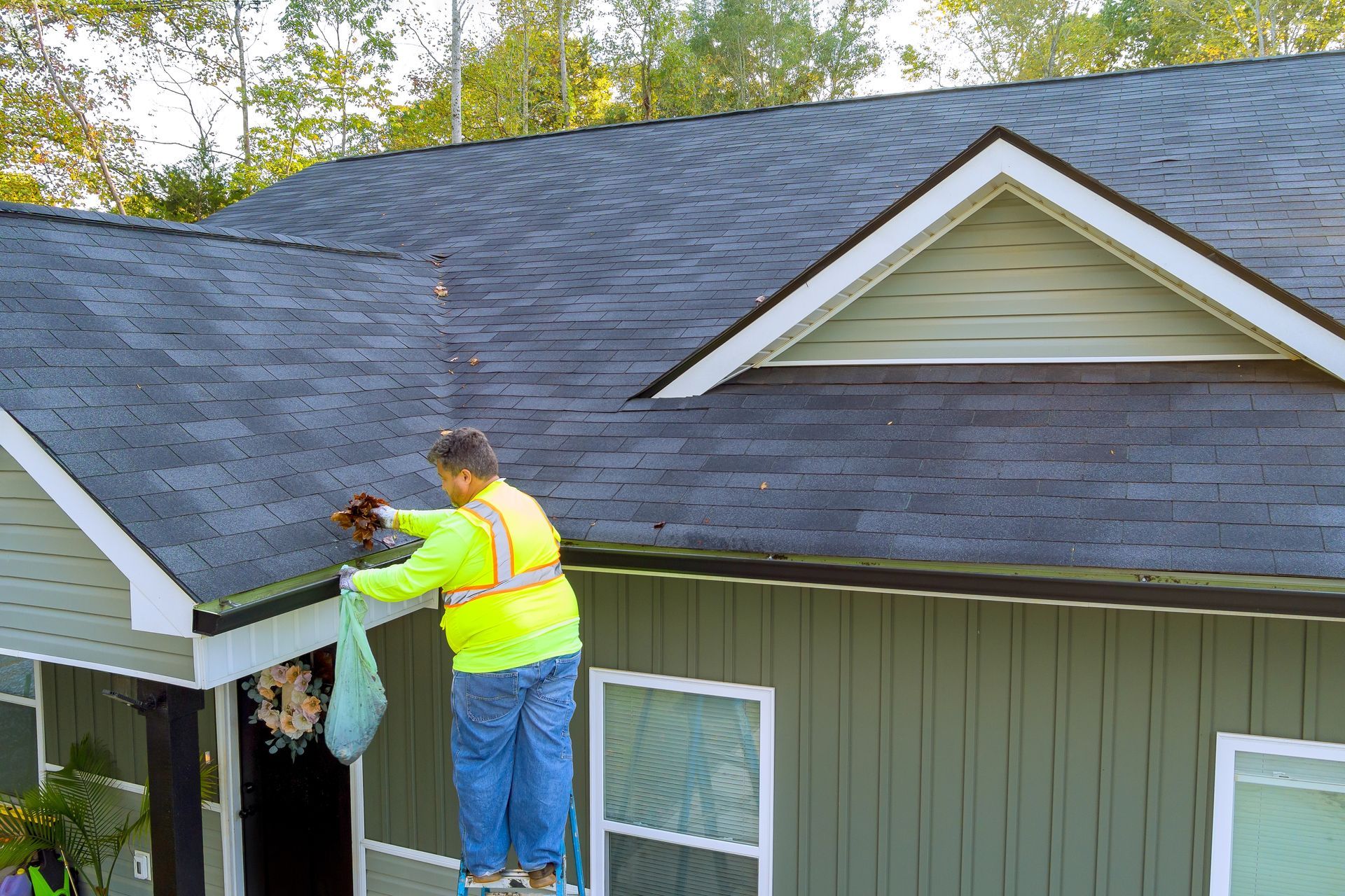 Man on ladder cleaning a roof gutter. He is wearing a safety vest and holding a bag.