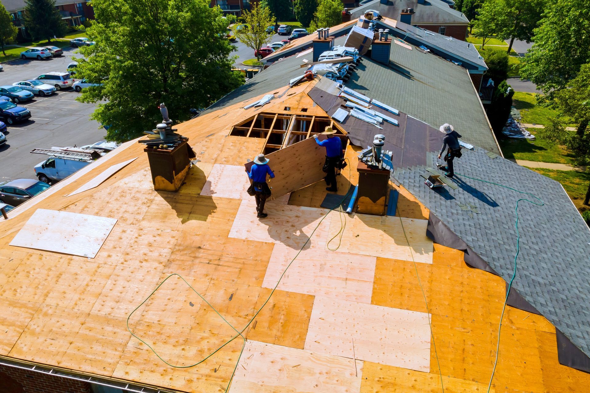 Roofers installing plywood on a residential roof, with chimneys and a parking lot visible.
