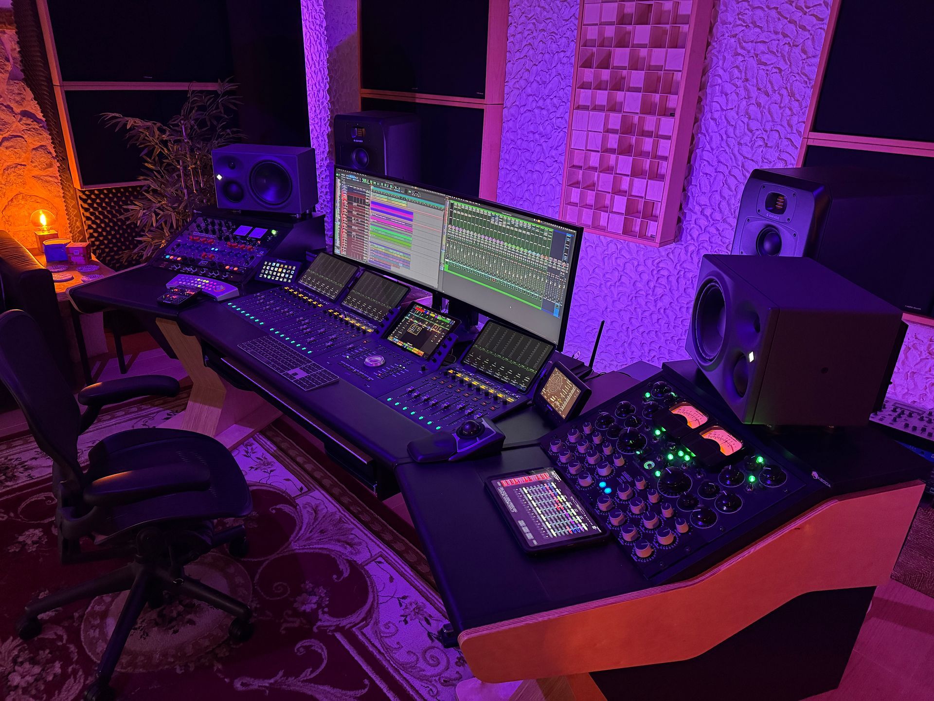 Recording studio desk with audio equipment, computer screen, and speakers; lit with purple light.