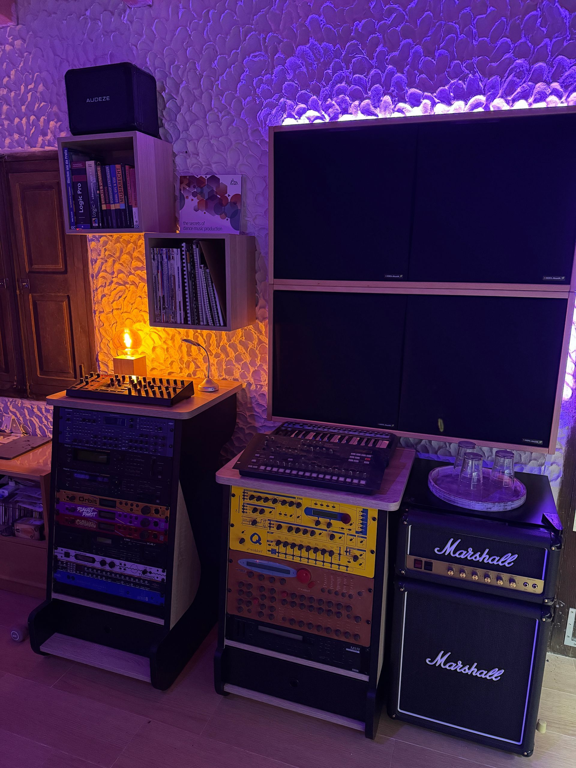 Music studio setup with stacks of equipment, a small amplifier, and speakers against a textured wall with purple lighting.