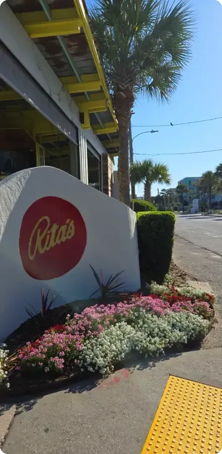 Sign for Rita's with colorful flowers in front of a building with a yellow roof in a coastal area.