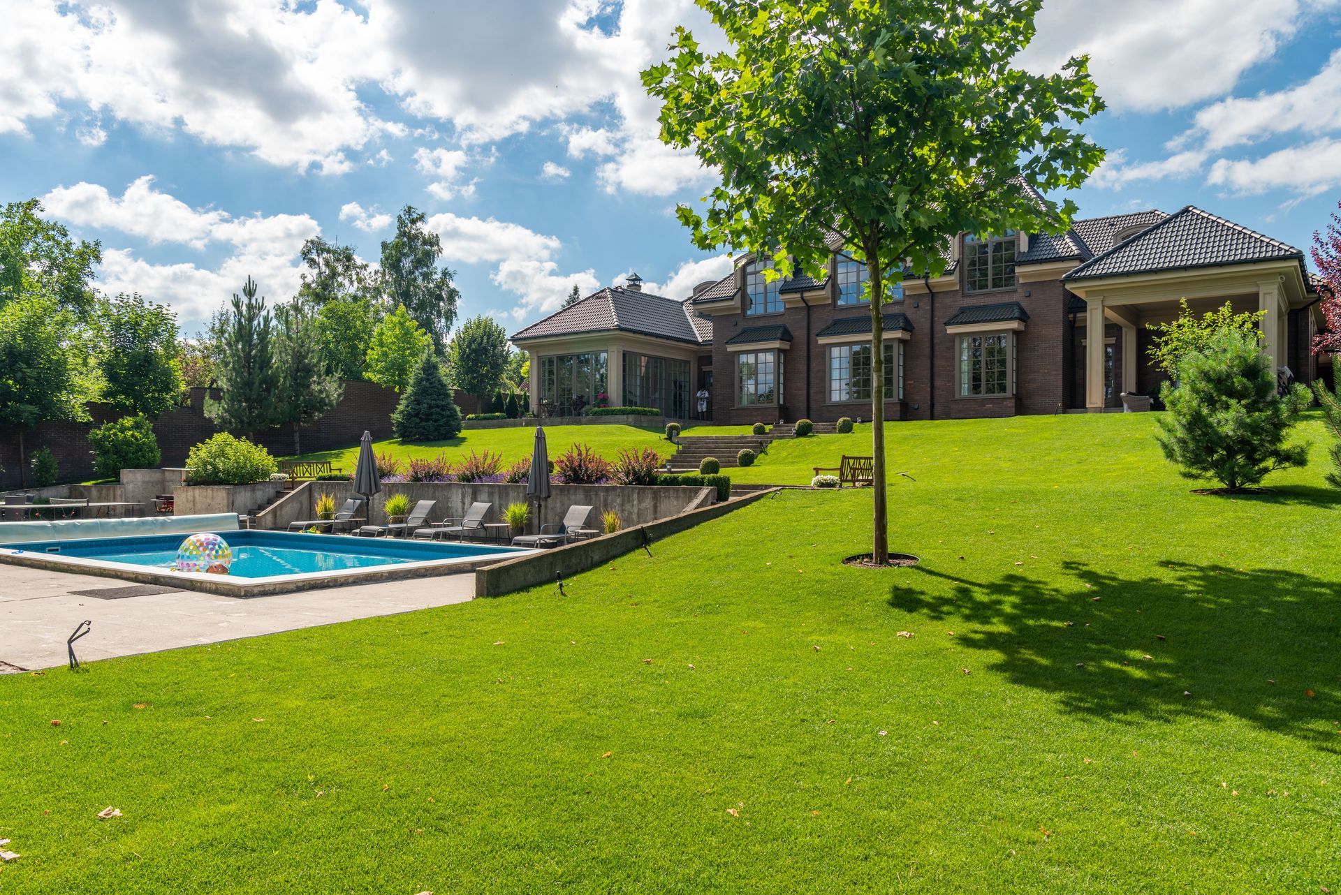 Large brick house with a green lawn, landscaped yard, and a swimming pool under a partly cloudy sky