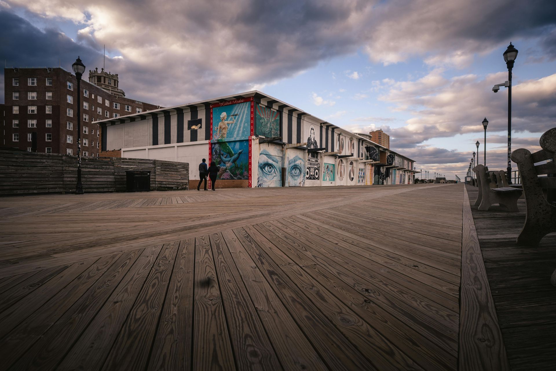 Boardwalk with building featuring colorful murals and cloudy sky. Two people walk.