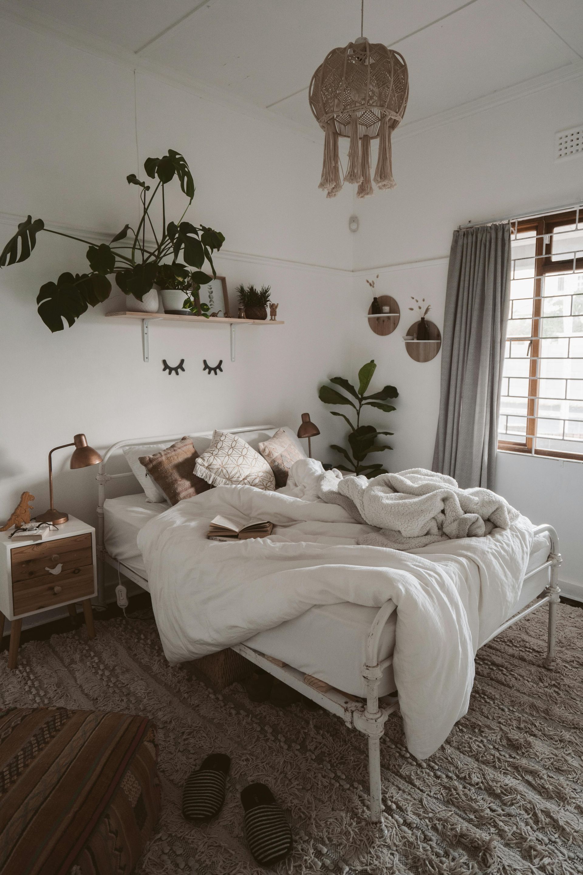 Cozy white bedroom with a bed, plants, and natural light. Woven chandelier and textured rug complete the bohemian vibe.