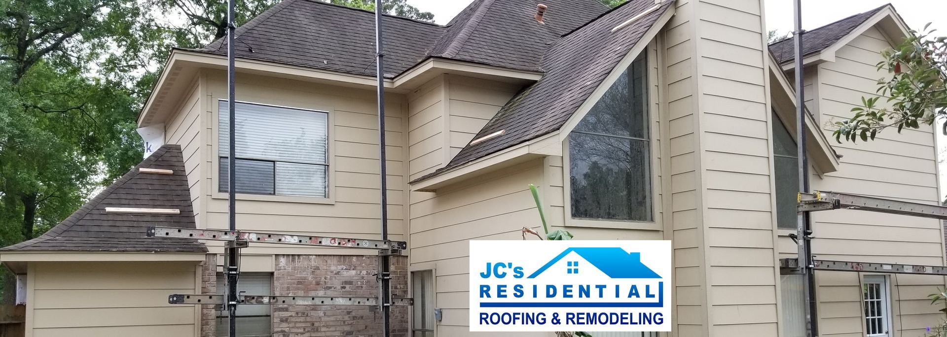 A multi-story house with dark brown roof under construction, beige siding, a large window, and the company logo.