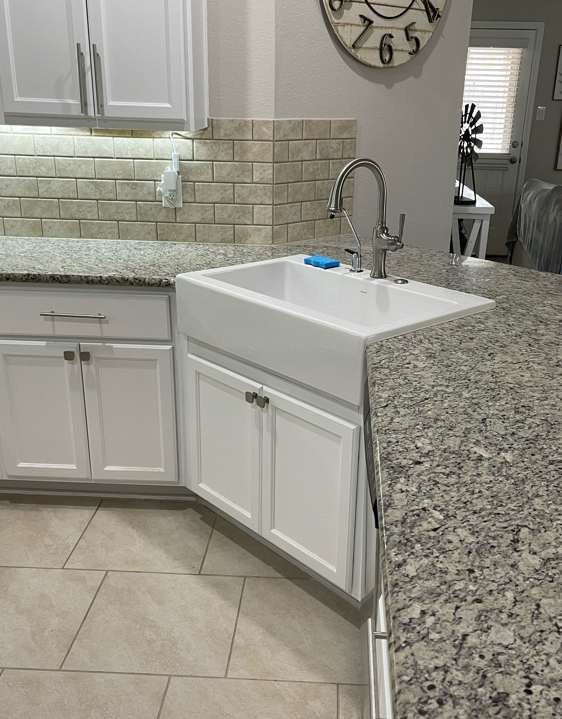 White kitchen sink with faucet, granite countertop, white cabinets, and tile backsplash.