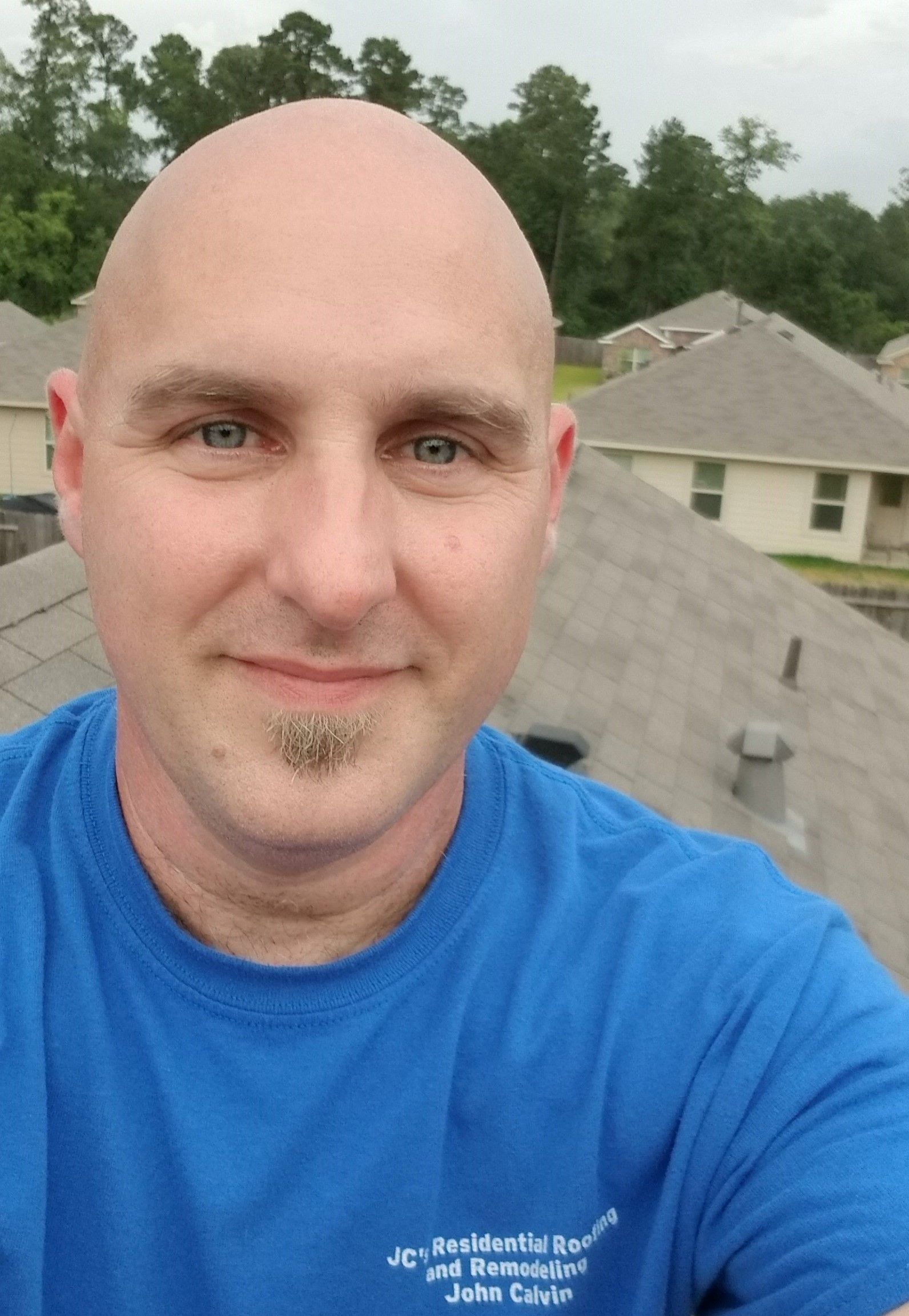 Bald man in blue shirt on a rooftop; smiling, trees and houses in the background.