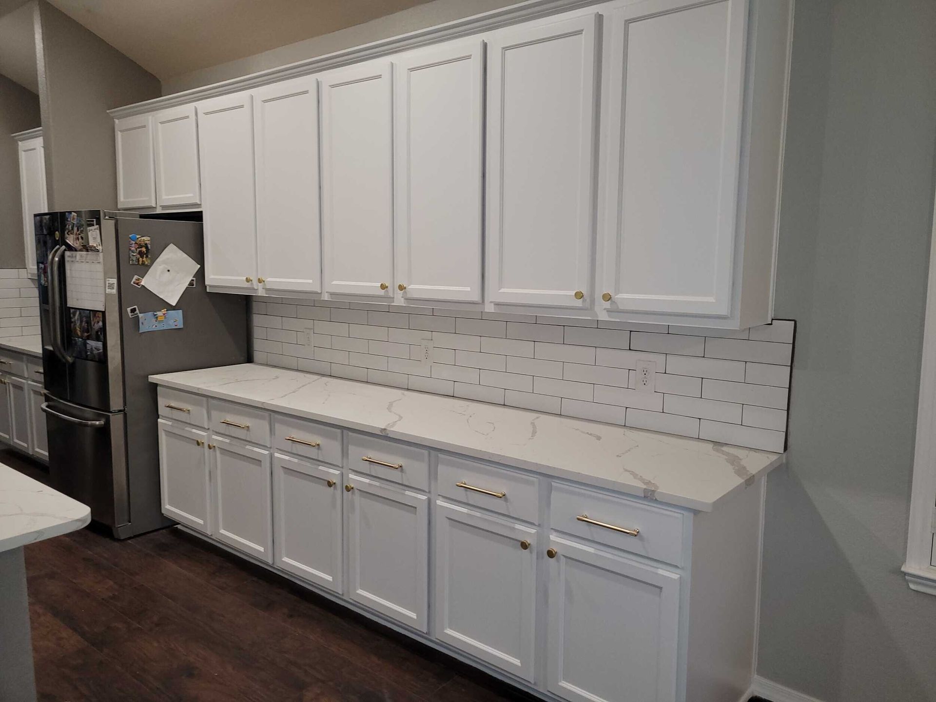 White kitchen cabinets, countertop, backsplash, and stainless steel refrigerator against a grey wall.