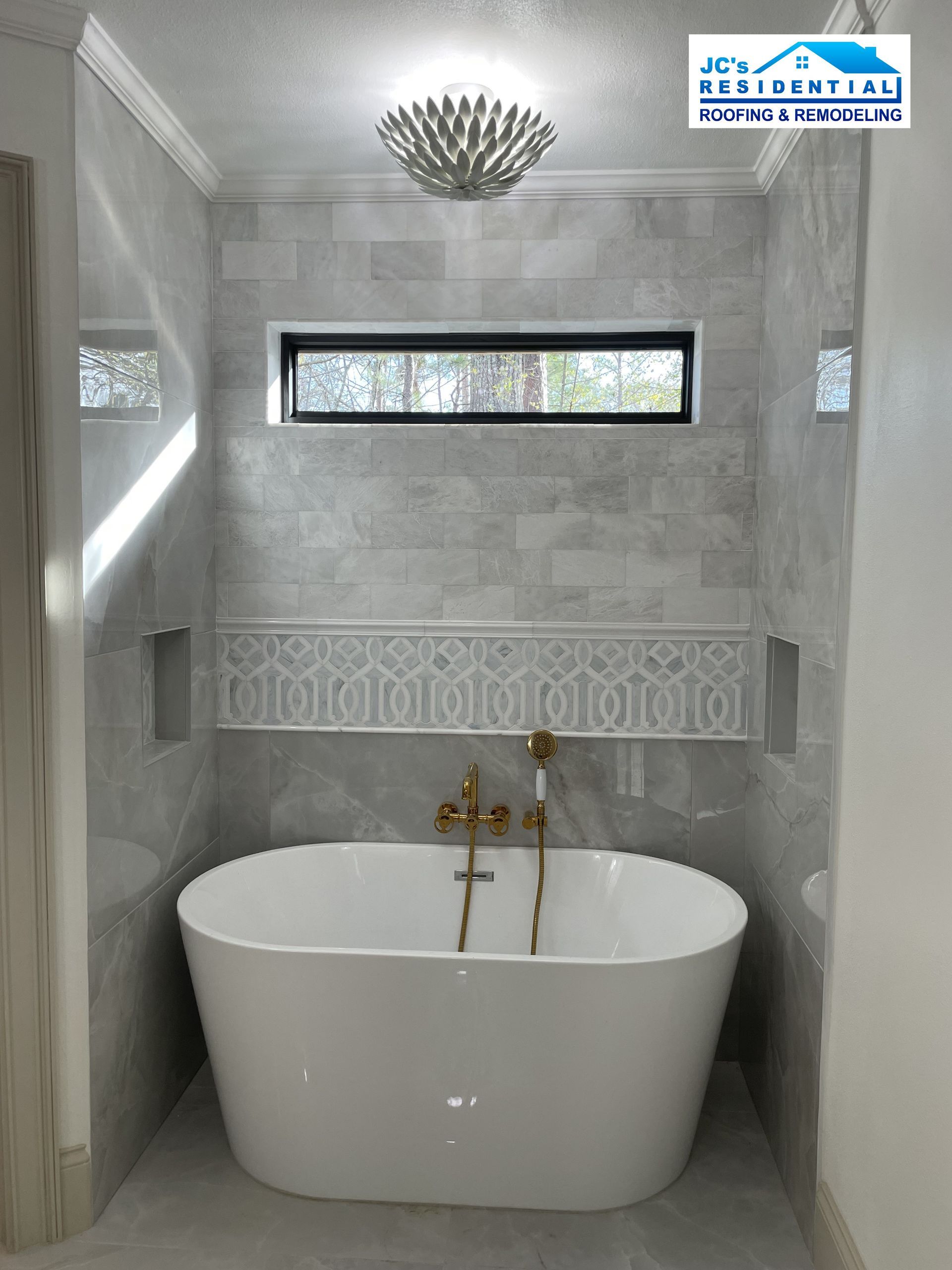 Bathroom with white tub, gold fixtures, gray tiled walls, and small window.