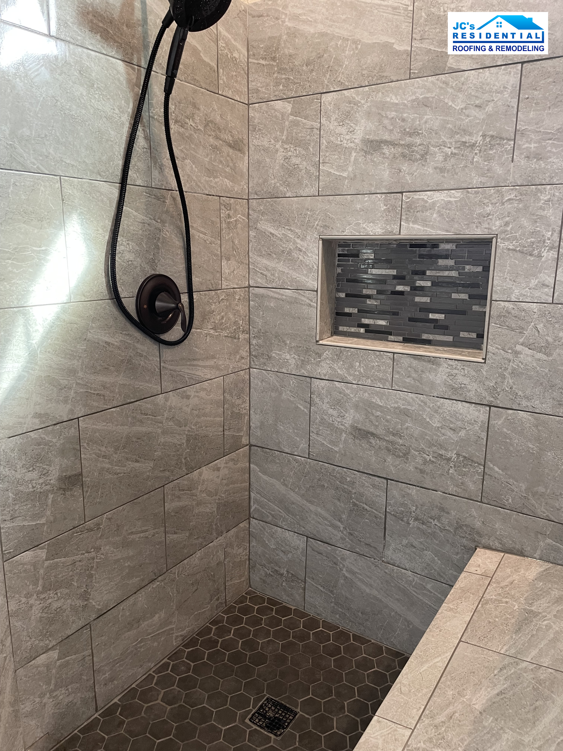 Shower with gray tile, a dark showerhead, and a tiled niche with small, dark mosaic tiles.