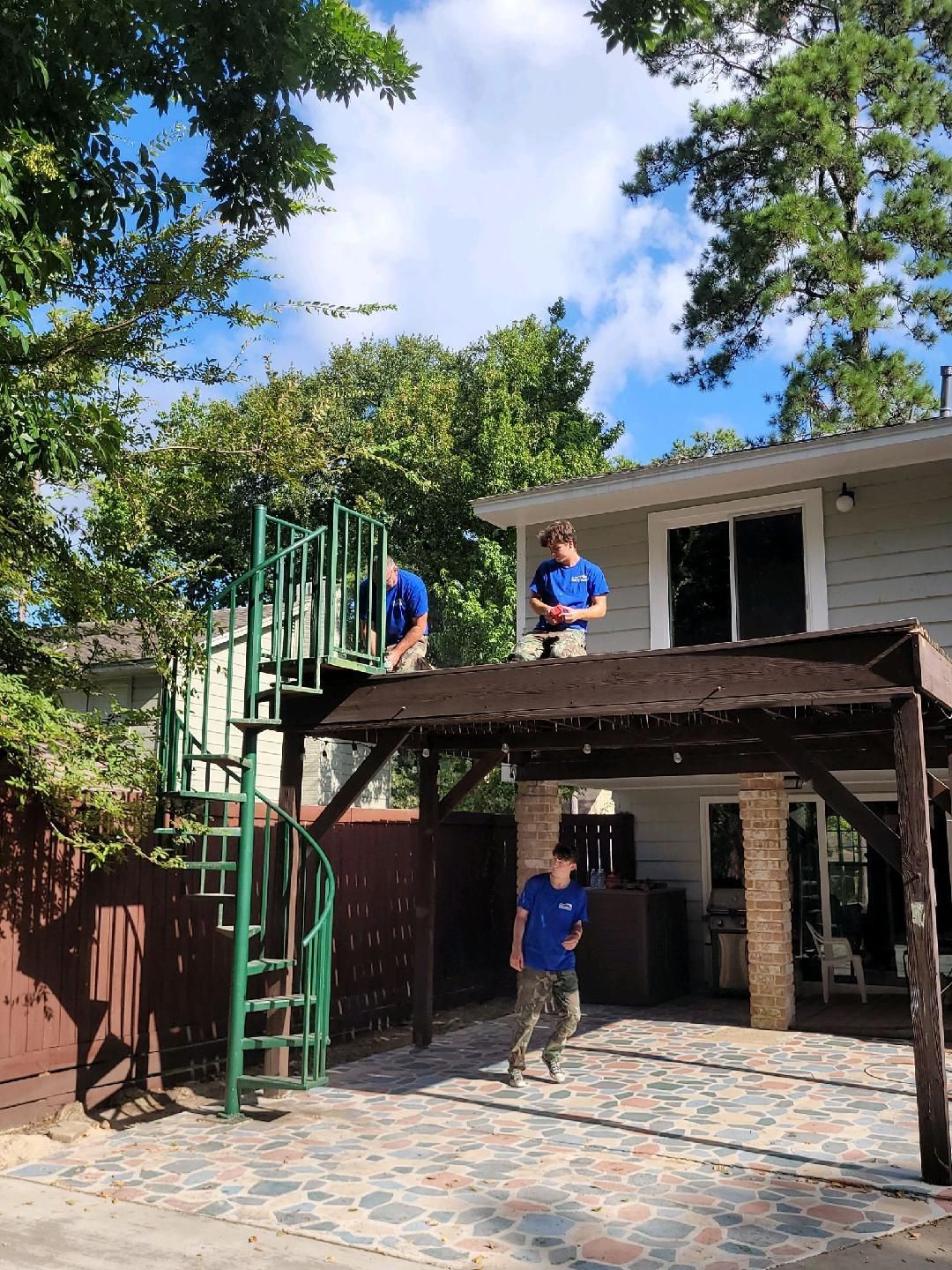 Three people on a rooftop patio; one on the ground. Green spiral staircase, trees, and blue sky.