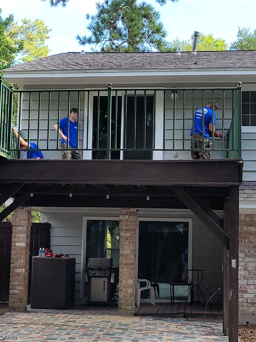 Three people painting a deck railing, wearing blue shirts, on a two-story deck with a gray house in the background.