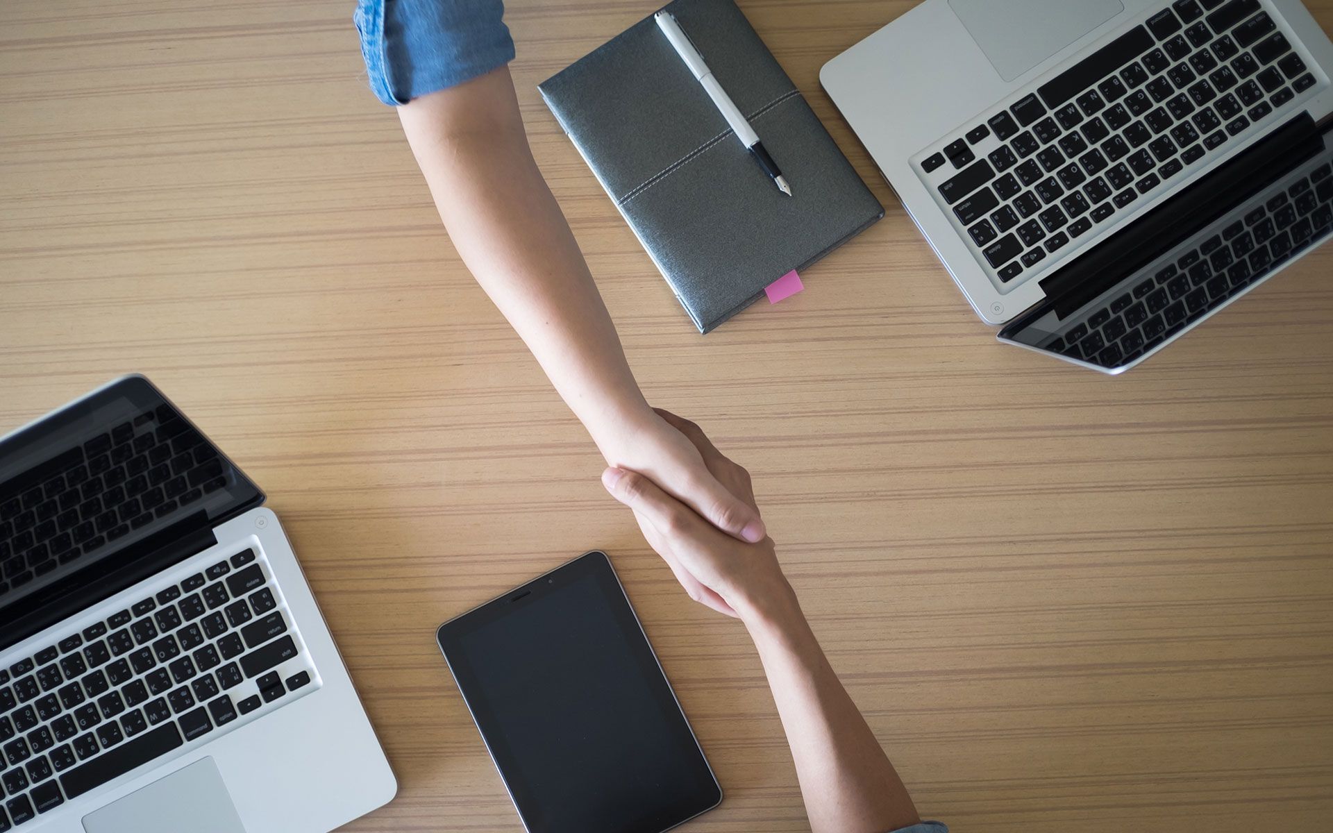 Two people are shaking hands over a wooden table with laptops.