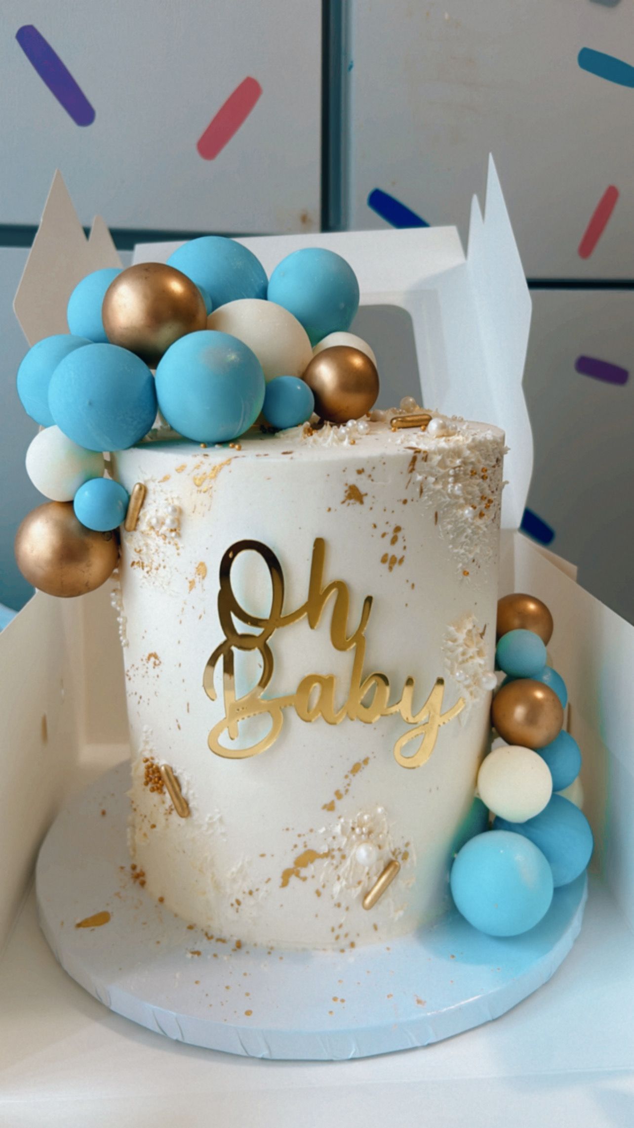 A white cake with blue and gold balls and the words oh baby on it
