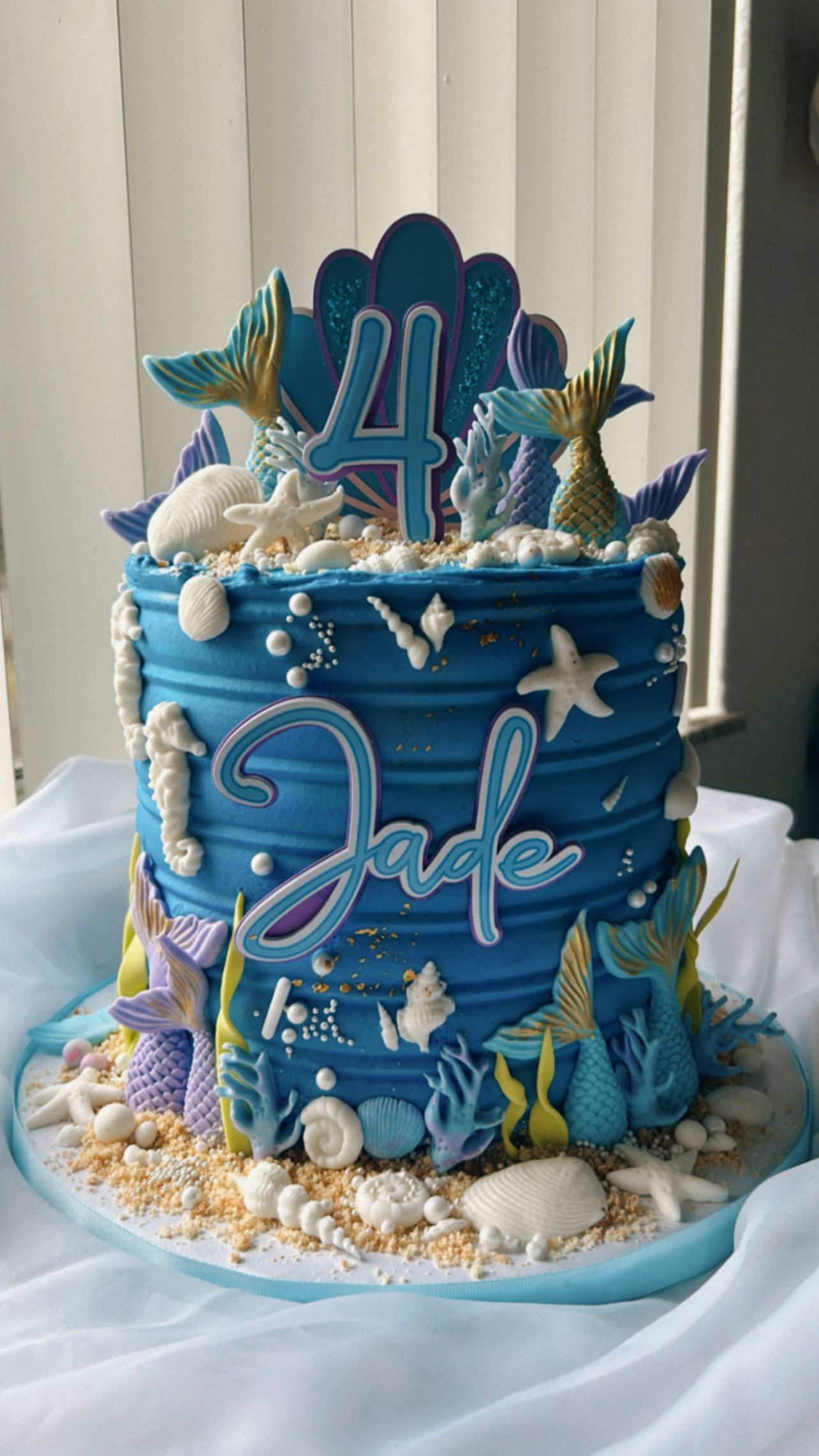 A blue cake with mermaids and seashells on it is sitting on a table.