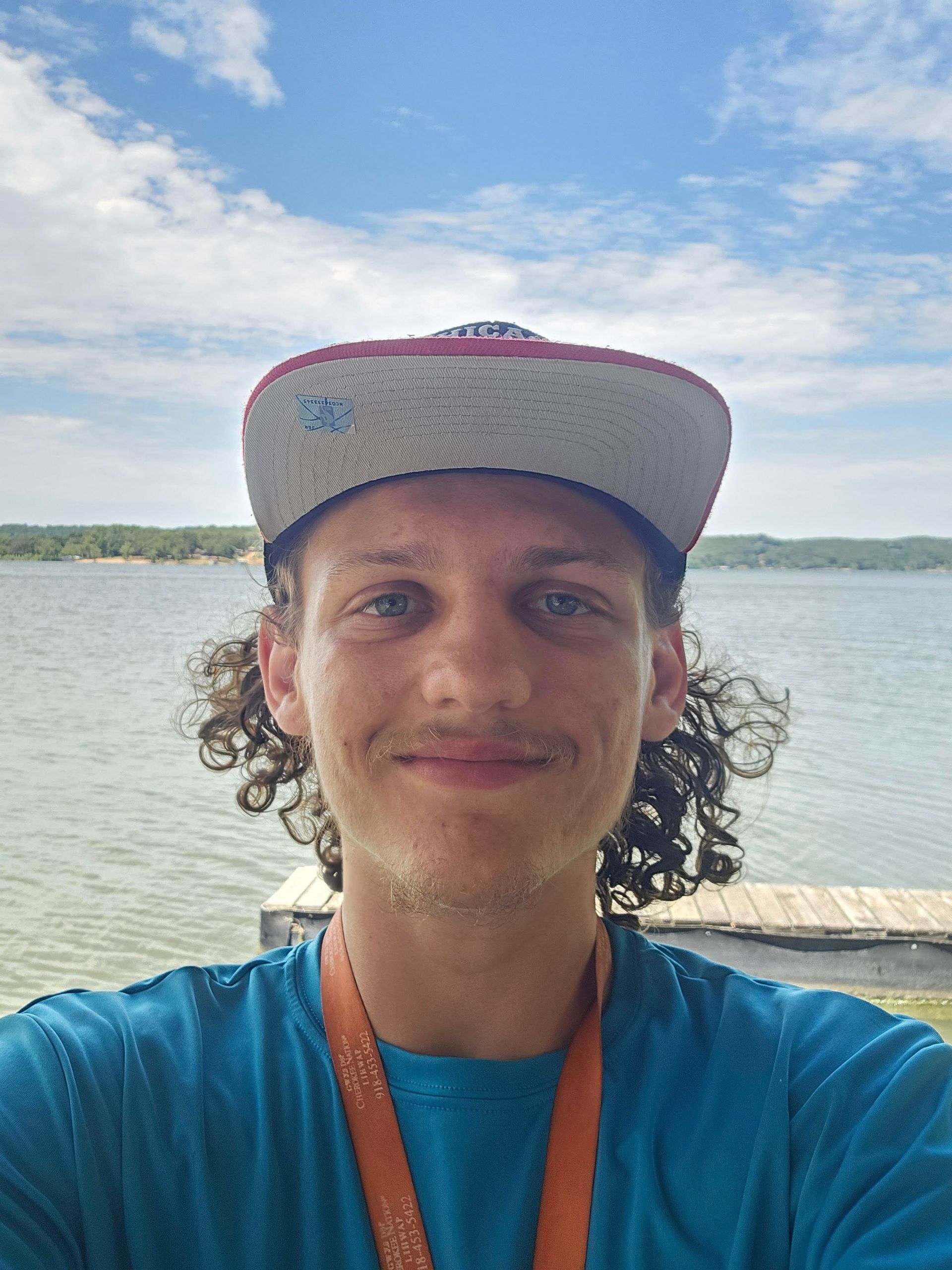 A young man wearing a hat is taking a selfie in front of a lake.