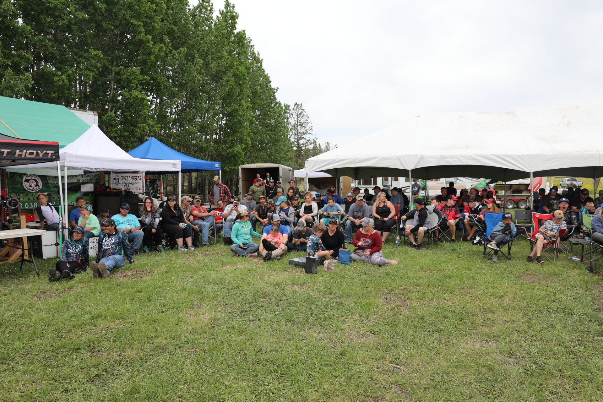 A large group of people are sitting in a grassy field under tents.
