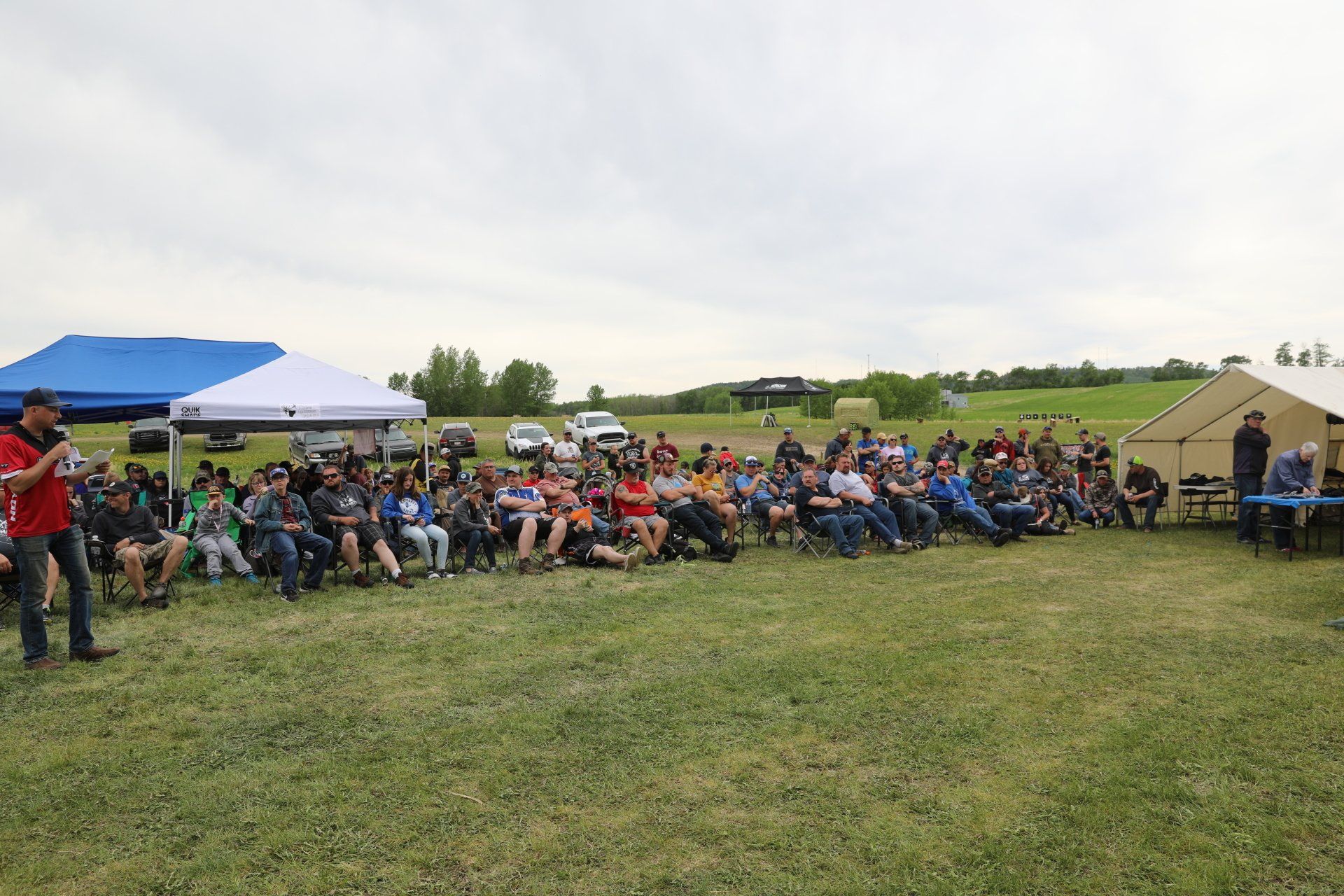 A man is standing in front of a large group of people sitting in chairs in a field.