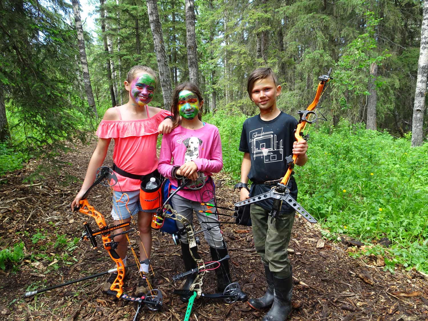 Three children are standing in the woods holding bows and arrows.