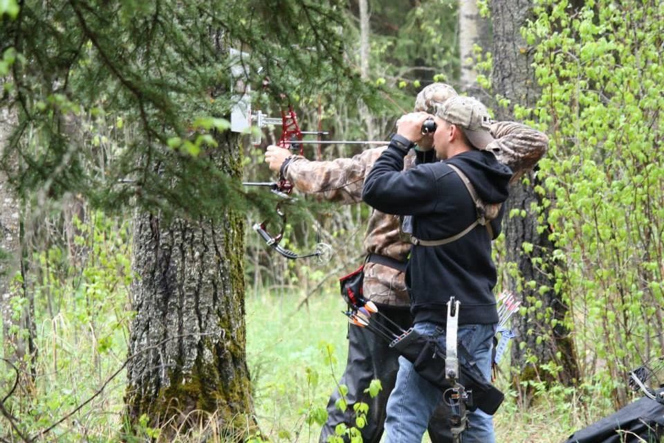 Two men are standing in the woods holding bows and arrows.