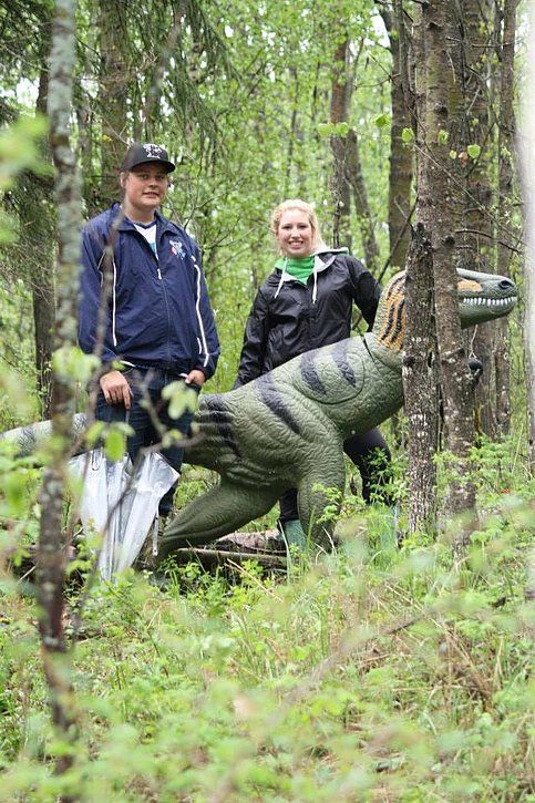 A man and a woman are standing next to a dinosaur statue in the woods.