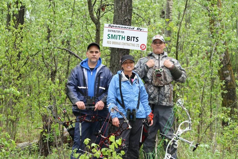 Three people are standing next to each other in the woods holding bows and arrows.