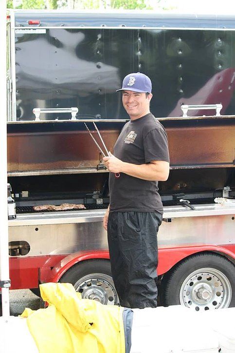 A man is standing in front of a food truck holding tongs.