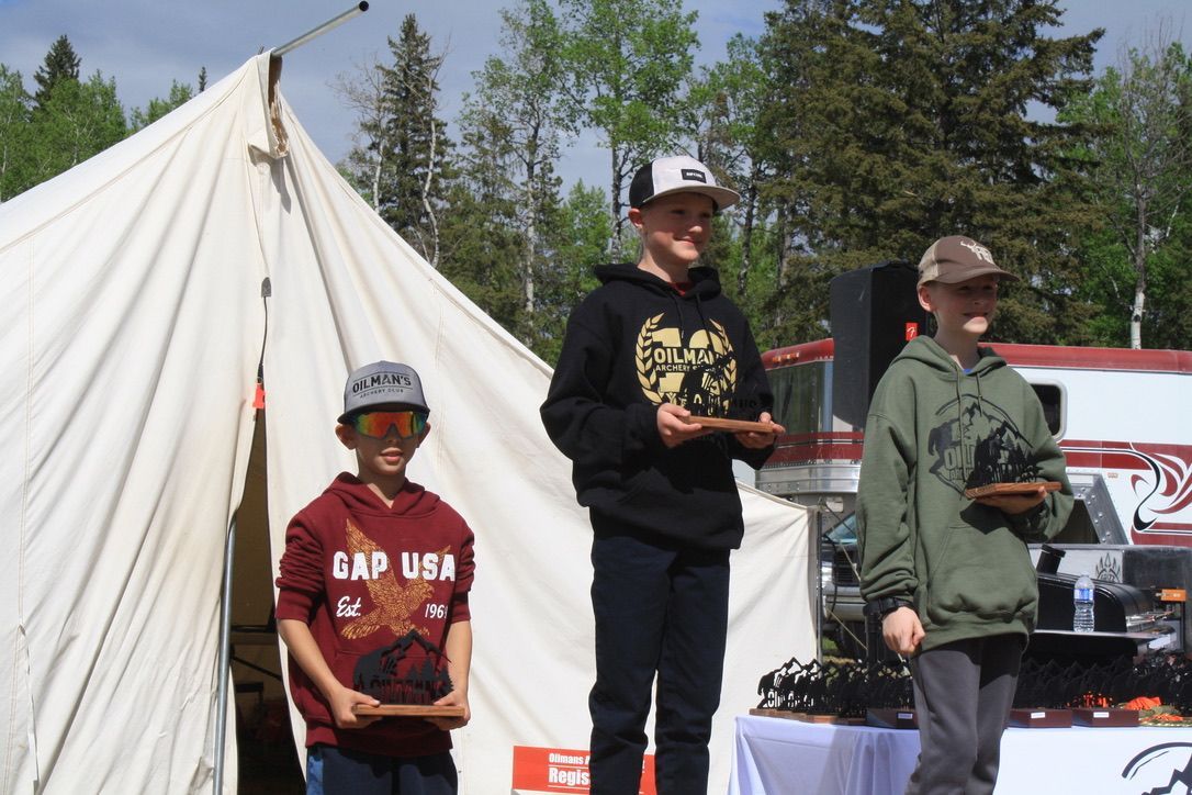 Three boys are standing in front of a tent holding trophies . one of the boys is wearing a gap usa shirt.
