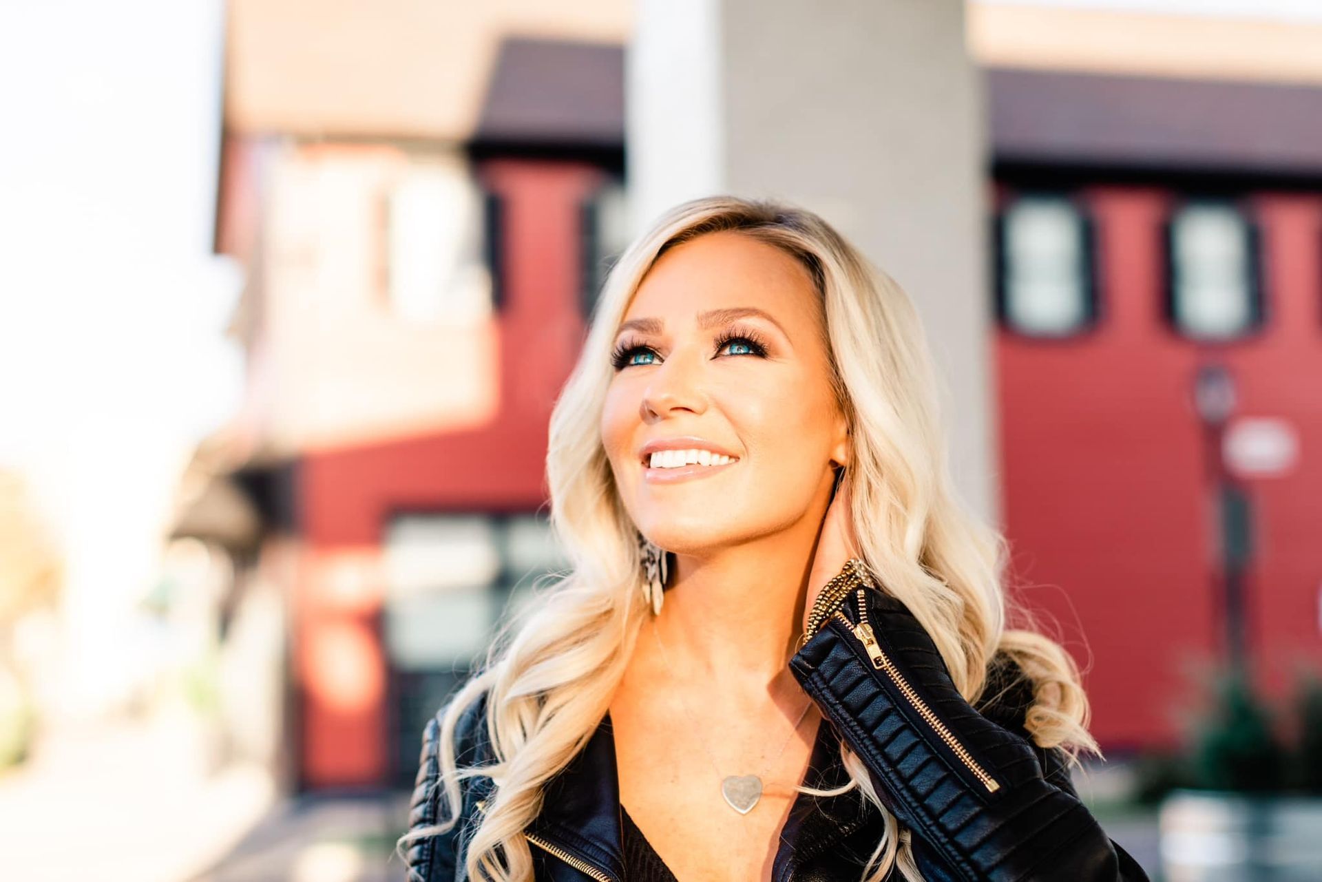 A woman in a black leather jacket is smiling in front of a red building.