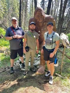 Three men are standing next to a statue of a bigfoot in the woods.