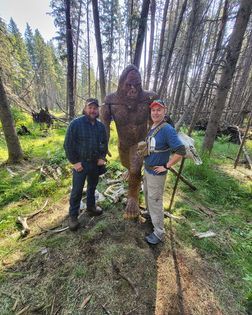 Two men are standing next to a statue of a bigfoot in the woods.
