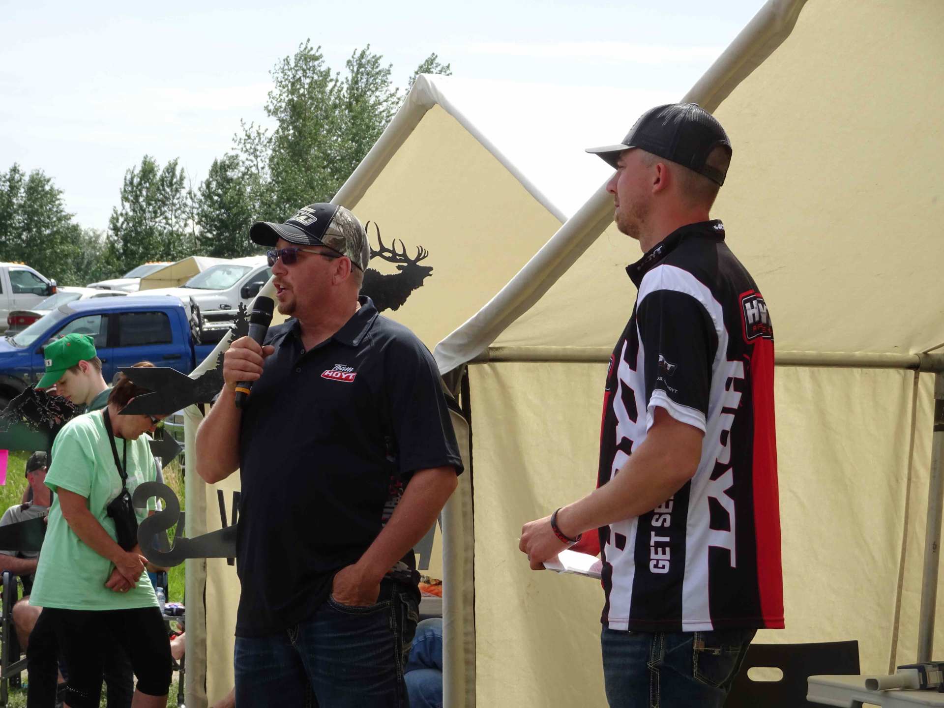 Two men are standing in front of a tent and one of them is holding a microphone
