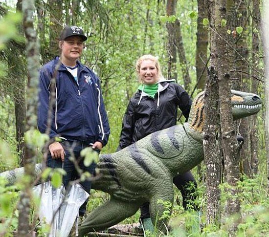 A man and a woman are standing next to a dinosaur statue in the woods.