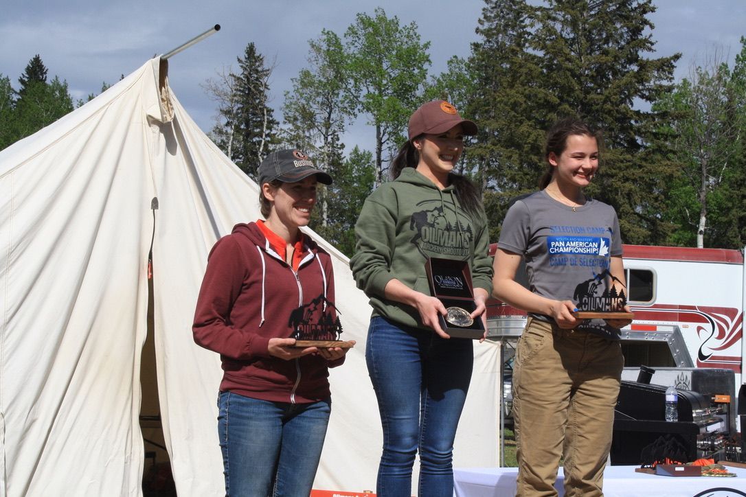 Three women are standing in front of a tent holding trophies.