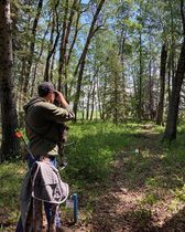 A man is standing in the middle of a forest looking through binoculars.