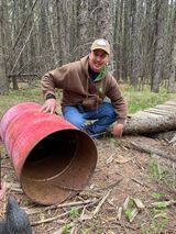 A man is kneeling next to a red barrel in the woods.