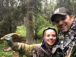 A man and a woman are standing next to a dinosaur in the woods.