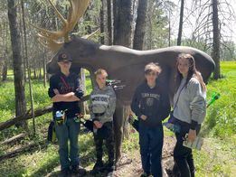 A group of people are standing in front of a moose statue in the woods.