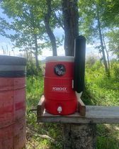 A red cooler is sitting on top of a wooden bench in the woods.