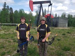 Two young men are standing in a field holding bows and arrows.