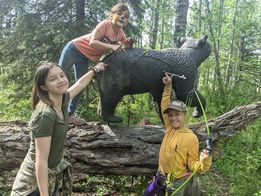 Two girls are standing next to a statue of a bear in the woods.